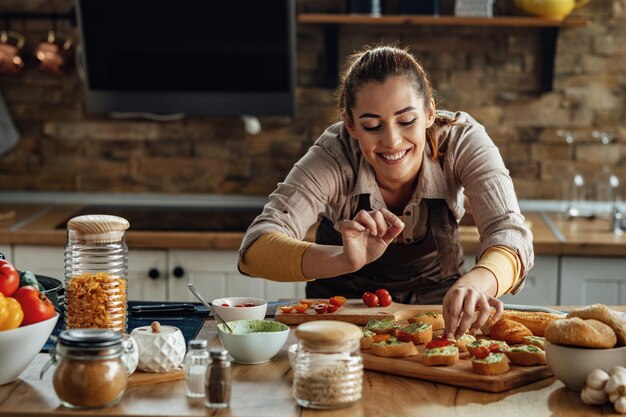 Junge lächelnde Frau, die Bruschetta mit gesunden Zutaten zubereitet, während sie Essen in der Küche zubereitet