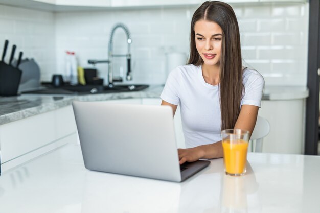 Junge lächelnde Frau, die Frühstück in der Küche verbindet, die mit einem Laptop verbindet und einen gesunden Orangensaft trinkt