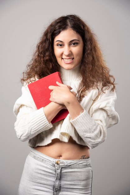 Junge lächelnde Frau, die rotes Buch auf einer grauen Wand hält. Hochwertiges Foto