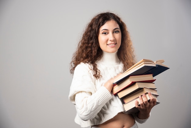 Junge lächelnde Frau, die viele Bücher auf einer grauen Wand trägt. Hochwertiges Foto