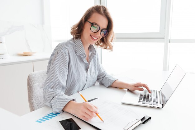 Junge lächelnde Frau in den Gläsern und im gestreiften Hemd, die mit Dokumenten und Computer arbeiten, während sie am Tisch in der hellen Küche sitzen