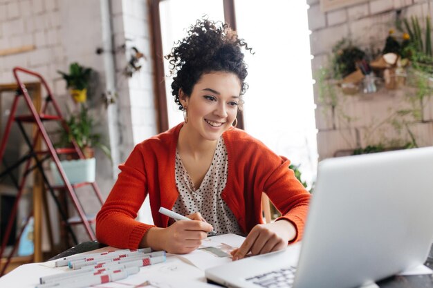 Junge lächelnde Frau mit dunklem, lockigem Haar, die am Tisch sitzt und glücklich am Laptop arbeitet, um Modeillustrationen zu zeichnen, während sie Zeit in einer modernen, gemütlichen Werkstatt mit großen Fenstern verbringt