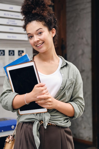 Kostenloses Foto junge lächelnde frau mit dunklem lockigem haar in khakifarbenem hemd, die lehrbücher und tablet in den händen hält und freudig in die kamera auf der straße der stadt schaut