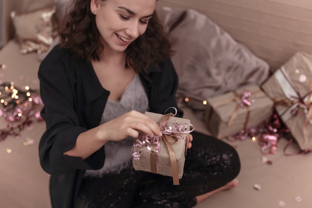 Junge lächelnde Frau packt ein Weihnachtsgeschenk aus, während sie zu Hause auf dem Bett sitzt.