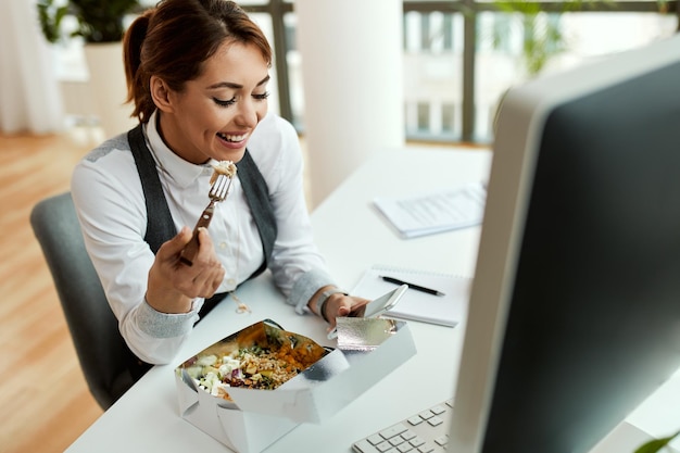 Junge lächelnde Geschäftsfrau, die beim Essen an ihrem Schreibtisch eine SMS auf dem Handy schreibt