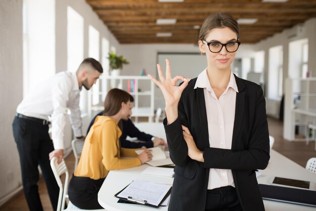 Junge lächelnde Geschäftsfrau mit Brille und Hemd, die verträumt in die Kamera blickt und eine Ok-Geste zeigt, während sie Zeit im Büro mit Kollegen im Hintergrund verbringt