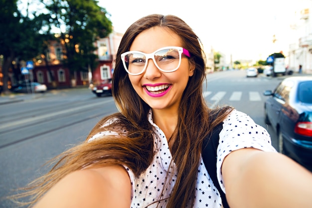 Junge lächelnde jugendlich glückliche Frau, die Selfie auf der Straße macht, ling Haare, helles Make-up und niedliche klare Brille, allein reisend, Spaß, positive Stimmung, Freude, Urlaub machend