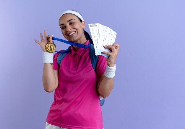 Junge lächelnde kaukasische sportliche Frau, die Rückentaschenstirnband und Armbänder trägt, hält goldene Medaille und Flugtickets lokalisiert auf lila Hintergrund mit Kopienraum