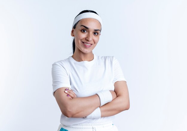 Junge lächelnde kaukasische sportliche Frau, die Stirnband und Armbänder trägt, steht mit verschränkten Armen lokalisiert auf weißem Raum mit Kopienraum