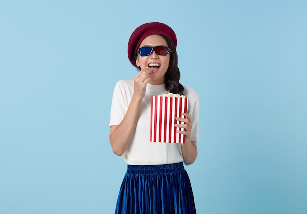Junge lächelnde, lustige, coole asiatische Frau mit 3D-Brille schaut sich einen Film an und hält einen Eimer Popcorn in der Hand