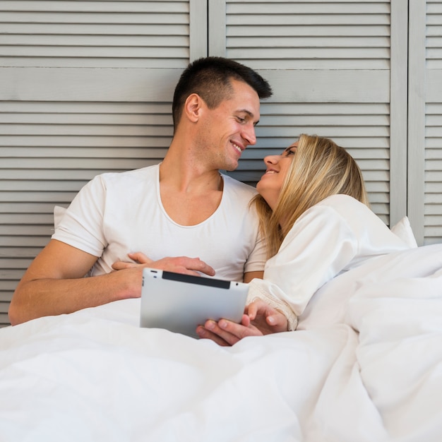 Junge lächelnde Paare mit Tablette unter Decke auf Bett