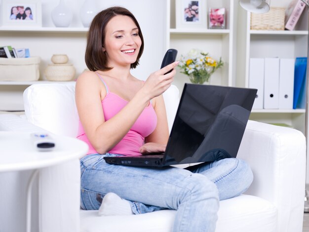Junge lächelnde schöne Frau, die auf dem Sofa mit Telefon und Laptop sitzt