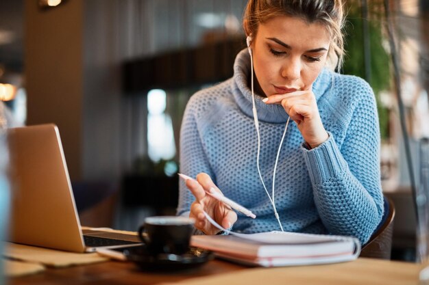 Junge lässige Geschäftsfrau, die in einem Café arbeitet und Papierkram durchgeht, während sie Musik über Kopfhörer hört