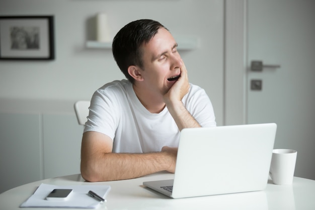 Junge langweilig, gähnen Mann, sitzt am weißen Schreibtisch in der Nähe von Laptop