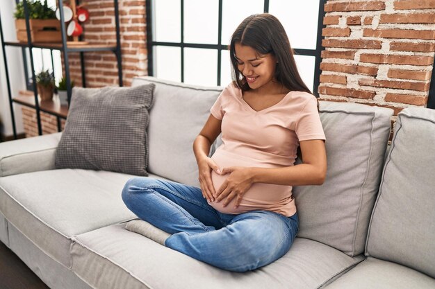 Junge lateinamerikanische Frau, die schwanger ist und den Bauch berührt, sitzt zu Hause auf dem Sofa