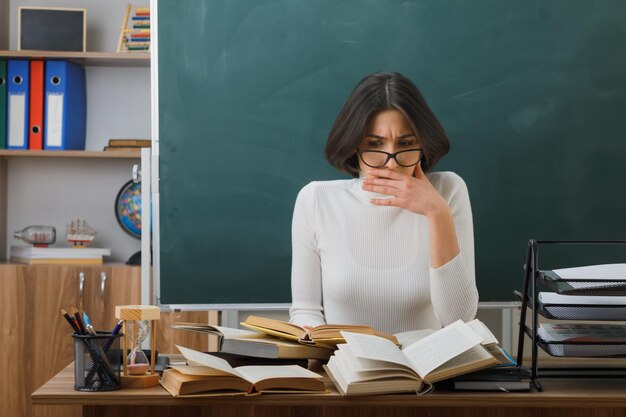 junge lehrerin, die am schreibtisch mit schulwerkzeugen im klassenzimmer sitzt