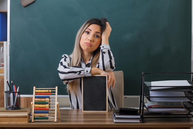 junge lehrerin, die an der schulbank vor der tafel im klassenzimmer sitzt und den unterricht erklärt, der eine kleine tafel hält und müde und überarbeitet in die kamera schaut