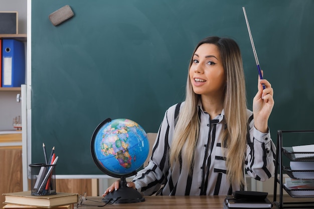 junge lehrerin, die an der schulbank vor der tafel im klassenzimmer sitzt und den unterricht erklärt, der globus und zeiger hält und selbstbewusst lächelnd aussieht