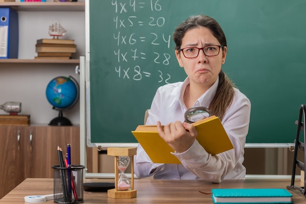 Junge Lehrerin, die eine Brille hält, die Buch betrachtet, das Seiten durch Vergrößerung betrachtet, verwirrt verwirrt wird, das an der Schulbank vor der Tafel im Klassenzimmer sitzt