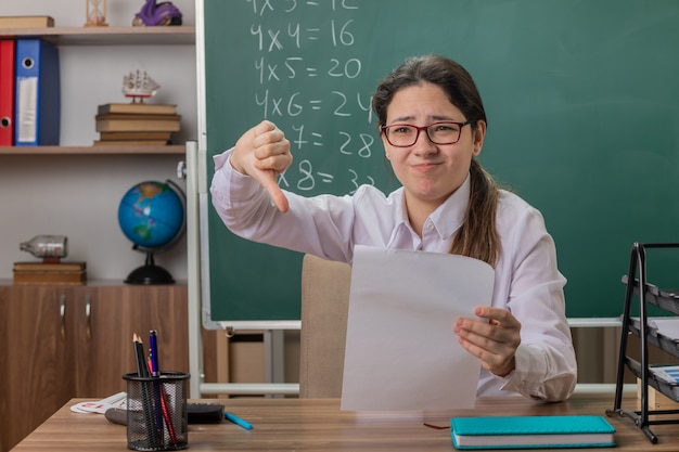 Junge Lehrerin, die eine Brille trägt, die an der Schulbank mit leeren Seiten sitzt, die verwirrt und unzufrieden aussehen und Daumen nach unten zeigen, die Hausarbeit vor Tafel im Klassenzimmer überprüfen