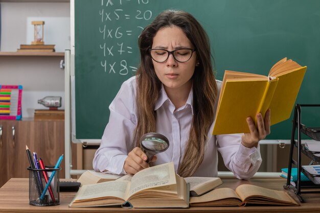 Junge Lehrerin, die eine Brille trägt, die Buch durch Lupe betrachtet, das fasziniert ist, das an der Schulbank vor der Tafel im Klassenzimmer sitzt