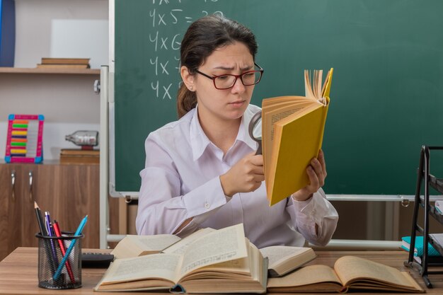 Junge Lehrerin, die eine Brille trägt, die Buch durch Lupe betrachtet, die verwirrt und unzufrieden ist, die an der Schulbank vor der Tafel im Klassenzimmer sitzt