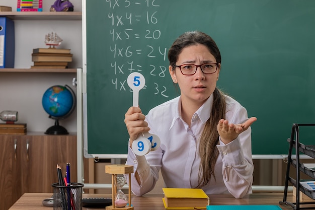 Junge Lehrerin, die eine Brille trägt, die Nummernschilder hält, die Lektion erklären, die mit dem Arm heraus verwirrt betrachtet, als fragend, wie sie an der Schulbank vor der Tafel im Klassenzimmer sitzt