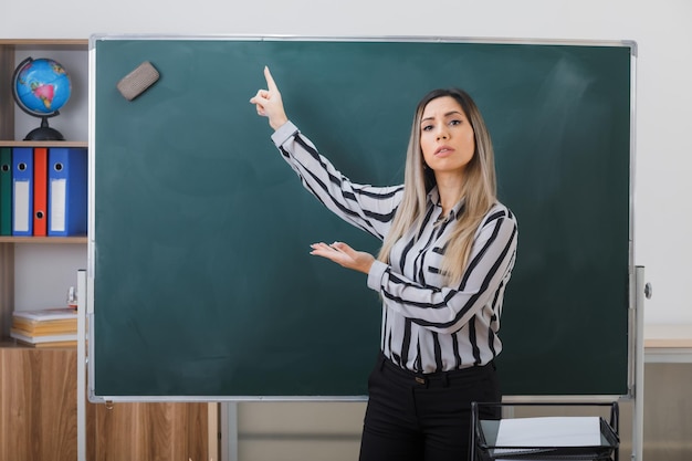 Junge lehrerin, die im klassenzimmer neben der tafel steht, erklärt den unterricht, zeigt mit dem zeigefinger auf die tafel und präsentiert sich mit dem arm und sieht selbstbewusst aus