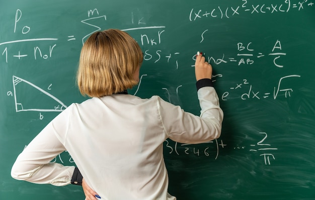 Kostenloses Foto junge lehrerin, die vor der tafel steht und etwas auf die tafel schreibt, mit gestrandet für tafel im klassenzimmer