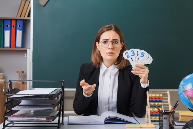 Junge Lehrerin mit Brille, die an der Schulbank vor der Tafel im Klassenzimmer sitzt und Nummernschilder hält, die den Unterricht erklären, sieht unzufrieden aus und hebt den Arm vor Unmut