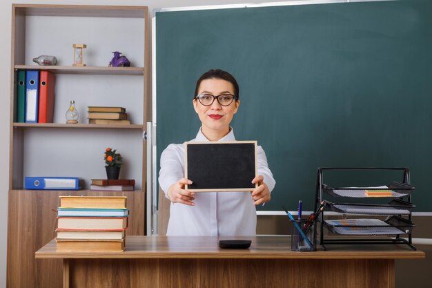 Junge Lehrerin mit Brille, die eine kleine Tafel zeigt, die selbstbewusst an der Schulbank vor der Tafel im Klassenzimmer sitzt