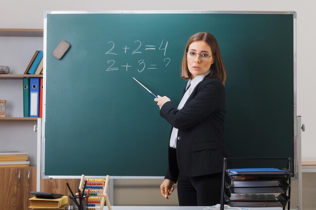 Junge Lehrerin mit Brille, die neben der Tafel im Klassenzimmer steht und den Unterricht erklärt, der auf die Tafel zeigt, wobei der Zeiger zuversichtlich aussieht