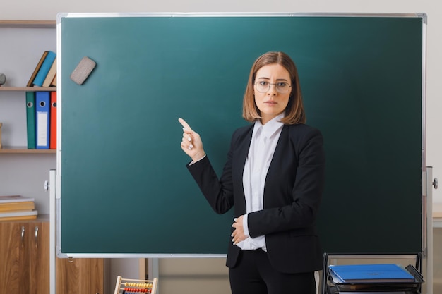 junge lehrerin mit brille, die neben der tafel im klassenzimmer steht und den unterricht erklärt, der mit dem zeigefinger auf die tafel zeigt und nervös und unzufrieden ist