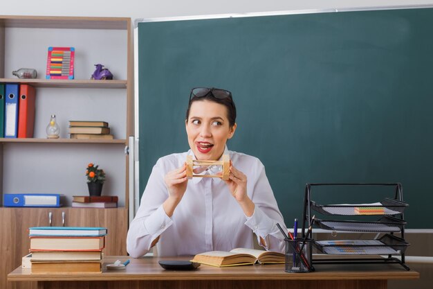 Junge Lehrerin mit Brille sitzt an der Schulbank mit Buch vor der Tafel im Klassenzimmer und hält eine Sanduhr mit verschmitztem Gesichtsausdruck