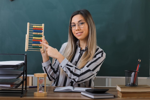 junge lehrerin mit brille sitzt an der schulbank vor der tafel im klassenzimmer und erklärt den unterricht mit abakus und lächelt selbstbewusst