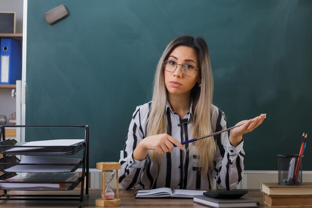 junge lehrerin mit brille sitzt an der schulbank vor der tafel im klassenzimmer und erklärt den unterricht mit dem zeiger, der mit ernstem gesicht wartet