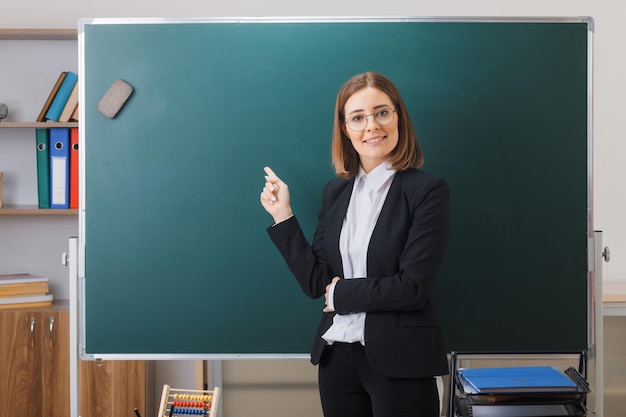 Junge Lehrerin mit Brille steht neben der Tafel im Klassenzimmer und erklärt den Unterricht, zeigt mit dem Zeigefinger auf die Tafel und lächelt selbstbewusst