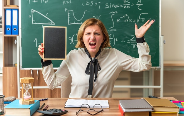 Kostenloses Foto junge lehrerin sitzt am tisch mit schulmaterial mit mini-tafel im klassenzimmer