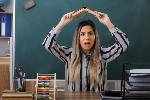 Kostenloses Foto junge lehrerin sitzt an der schulbank vor der tafel im klassenzimmer und hält buch über den kopf und sieht besorgt und verwirrt aus