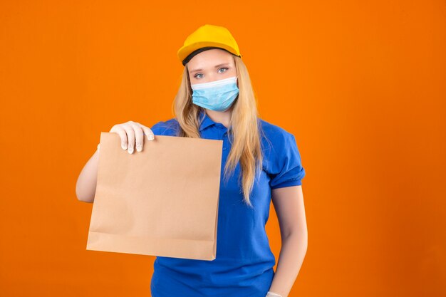 Junge Lieferfrau, die blaues Poloshirt und gelbe Kappe in der medizinischen Schutzmaske trägt, die Papierpaket in den Händen schaut, die über lokalem dunkelgelbem Hintergrund zuversichtlich schauen
