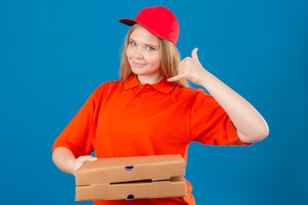 Kostenloses Foto junge lieferfrau, die orange poloshirt und rote kappe hält, die pizzakästen hält, die mich geste lächelnd freundlich über lokalisiertem blauem hintergrund halten