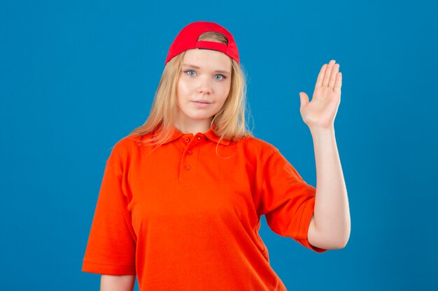 Junge Lieferfrau, die orange Poloshirt und rote Kappe lächelnd freundlich winkende Hand trägt, die Sie begrüßt und begrüßt oder auf Wiedersehen über isoliertem blauem Hintergrund sagt