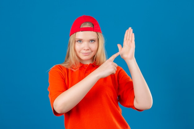 Junge Lieferfrau, die orange Poloshirt und rote Kappe zeigt, die Hand des offenen Kumpels betrachtet Kamera mit Lächeln auf Gesicht über lokalisiertem blauem Hintergrund zeigt