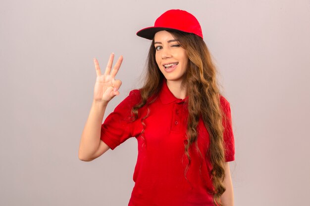 Junge Lieferfrau, die rotes Poloshirt und die Kappe zwinkert, die ok Zeichen über lokalisiertem weißem Hintergrund tun
