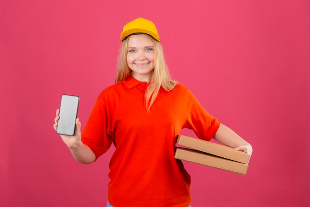 Junge Lieferfrau, die rotes Poloshirt und gelbe Kappe hält, die Pizzakästen hält, die Handy lächelnd freundlich über lokalisiertem rosa Hintergrund zeigen