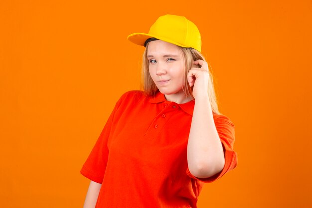 Junge Lieferfrau, die rotes Poloshirt und gelbe Kappe trägt, die Zweifel und mit verwirrendem Gesichtsausdruck über lokalisiertem orangefarbenem Hintergrund haben