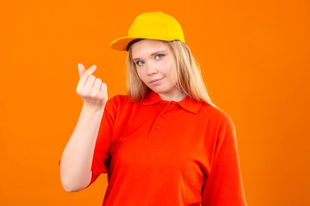 Junge Lieferfrau, die rotes Poloshirt und gelbe Kappe tut, die Geldgeste tut, die über lokalisierten orange Hintergrund lächelt