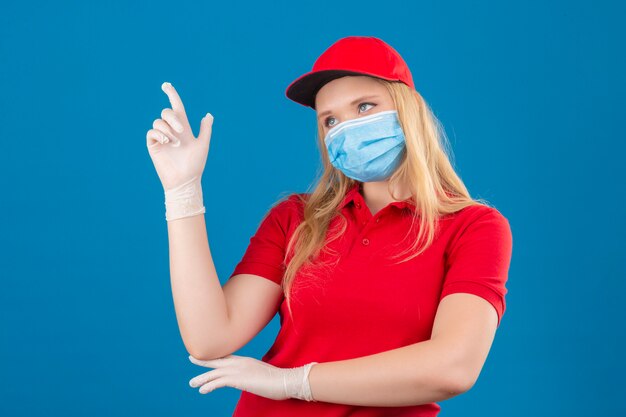 Junge Lieferfrau, die rotes Poloshirt und Kappe in der medizinischen Schutzmaske trägt, die mit dem Zeigefinger eine große Idee über lokalisiertem blauem Hintergrund zeigt