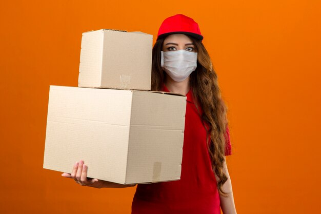 Junge Lieferfrau, die rotes Poloshirt und Kappe in der medizinischen Schutzmaske trägt, die mit Pappkartons steht, die über lokalisierten orange Hintergrund zuversichtlich schauen