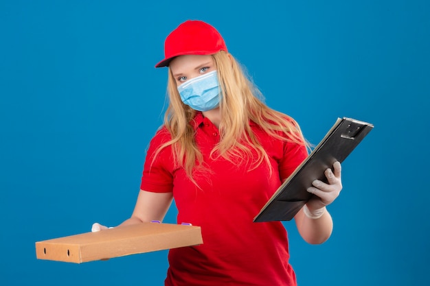 Junge Lieferfrau, die rotes Poloshirt und Kappe in der medizinischen Schutzmaske trägt, die mit Pizzakiste und Zwischenablage betrachtet Kamera mit ernstem Gesicht über lokalisiertem Hintergrund steht
